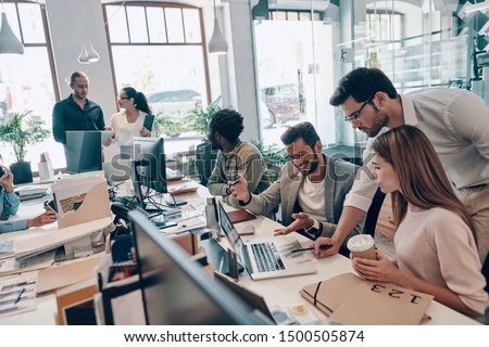 Group of young modern people in smart casual wear communicating and using modern technologies while working in the office              Royalty-Free Stock Photo #1500505874