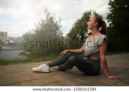Young woman runner resting after workout session on sunny morning. Female fitness model sitting on street along pond in city. Female jogger taking a break from running workout