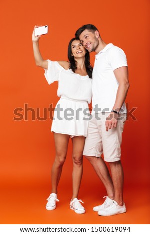 Full length of an attractive smiling young couple standing isolated over red background, taking a selfie