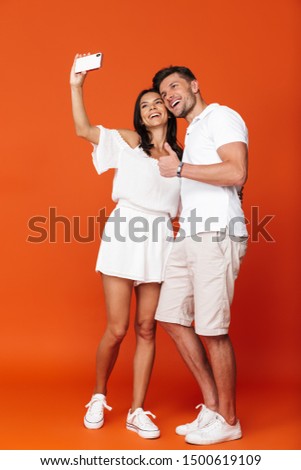 Full length of an attractive smiling young couple standing isolated over red background, taking a selfie