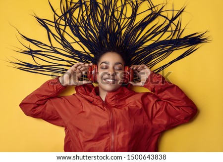 Portrait of beautiful african brunette teenage girl with dreadlocks wearing a red windbreaker listening music via wireless headphones isolated on vivid studio background