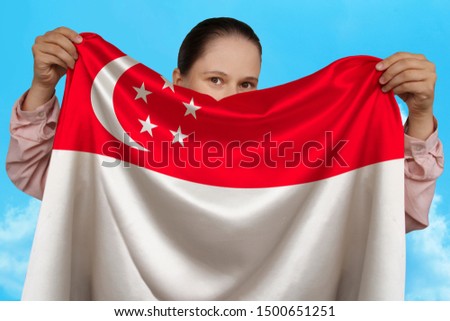 young girl holding in both hands the national flag of singapore on a beautiful shiny silk against a blue sky, concept state, travel, immigration, horizontal, close-up, copy space
