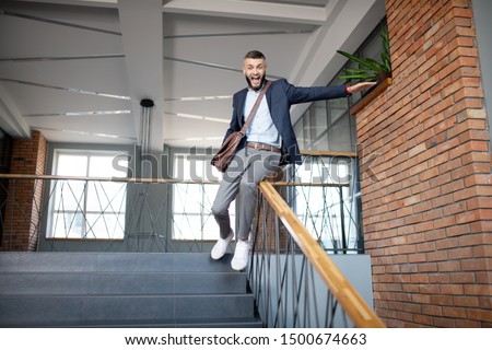 Sliding on banister. Cheerful bearded dark-haired man having fun while sliding on the banister Royalty-Free Stock Photo #1500674663