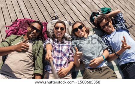 travel, tourism and people concept - group of friends or tourists with backpacks lying on wooden terrace and taking picture by selfie stick