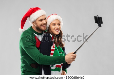 christmas, technology and holidays concept - happy couple in santa hats taking picture by smartphone on selfie stick at ugly sweater party