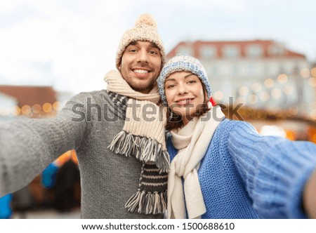people and winter holidays concept - happy couple in knitted hats and scarves taking selfie over christmas market in old town of tallinn city background