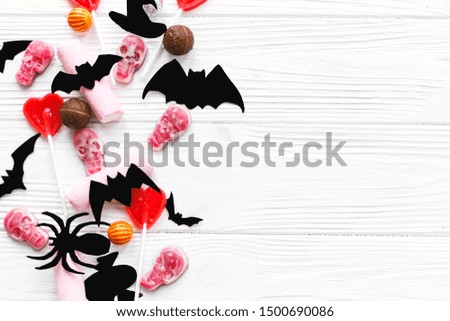 Halloween sweets flat lay. Halloween candy border with skulls, black bats, ghost, spider paper decorations on white wooden background, copy space. Trick or treat concept