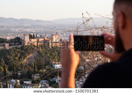 Alhambra palace and Granada cityscape being photographed by unfocused blurry bearded white man using a brandless phone camera to share in social media a picture taken from the Albaizin area.