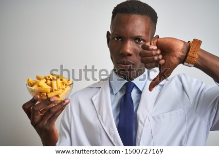 African american doctor man holding bowl with macaroni pasta over isolated white background with angry face, negative sign showing dislike with thumbs down, rejection concept