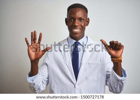 Young african american doctor man wearing coat standing over isolated white background showing and pointing up with fingers number six while smiling confident and happy.