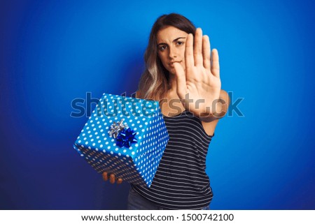 Young woman holding birthday present over blue isolated background with open hand doing stop sign with serious and confident expression, defense gesture