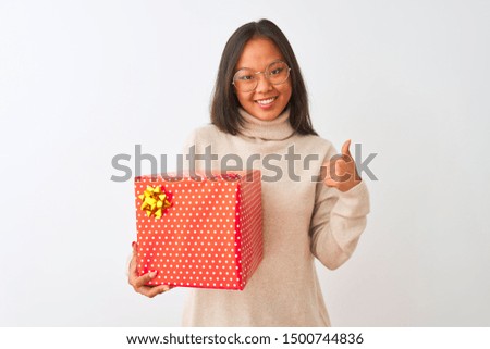 Young chinese woman wearing glasses holding birthday gift over isolated white background happy with big smile doing ok sign, thumb up with fingers, excellent sign