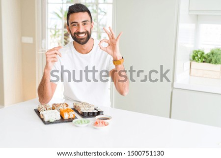 Handsome hispanic man eating asian sushi using chopsticks doing ok sign with fingers, excellent symbol