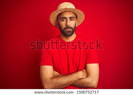 Young handsome indian man wearing t-shirt and hat over isolated red background skeptic and nervous, disapproving expression on face with crossed arms. Negative person.