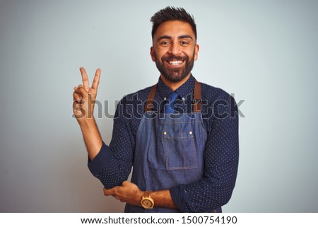 Young indian employee man wearing apron uniform standing over isolated white background smiling with happy face winking at the camera doing victory sign. Number two.