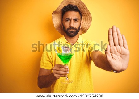 Indian man on vacation drinking cocktail wearing summer hat over isolated yellow background with open hand doing stop sign with serious and confident expression, defense gesture