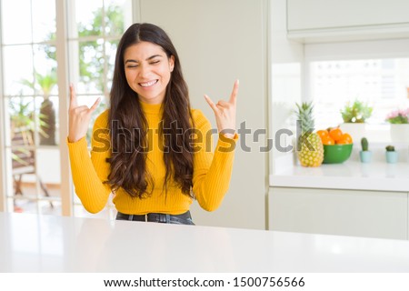 Young beautiful woman at home on white table shouting with crazy expression doing rock symbol with hands up. Music star. Heavy concept.
