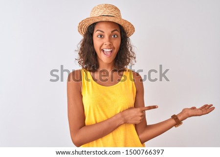 Young brazilian woman wearing yellow t-shirt and summer hat over isolated white background amazed and smiling to the camera while presenting with hand and pointing with finger.