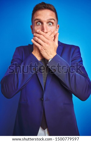 Vertical angle picture of young handsome business man over blue isolated background shocked covering mouth with hands for mistake. Secret concept.