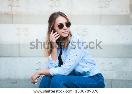 Image of beautiful stylish woman sitting on street stairs with legs crossed on summer day and holding mobile phone