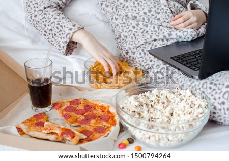 Unrecognized woman eating unhealthy food and crying. Caucasian teenage girl in cute warm pajamas sitting in the bed and watching movie on laptop. Unhealthy overeating lifestyle concept. Royalty-Free Stock Photo #1500794264