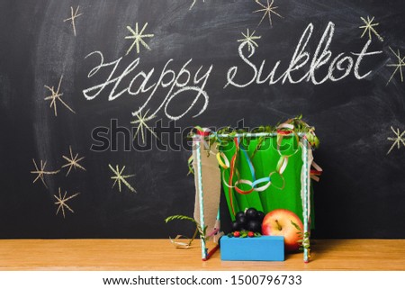 A hut made of paper covered with leaves and inscription on chalkboard Happy Sukkot.