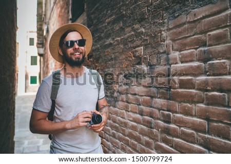 Male travel blogger spending vacations in Venice, Italy walkinbg on the narrow old town street with vintage film camera in hands. Bearded photographer standing outdoors looking for a good frame.  