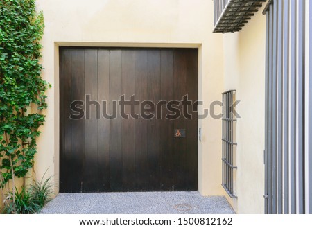 Dark brown wooden entrance door for wheelchairs with red sign in a small stoned alley with white walls and green plants running on one wall and fenced windows on the other.