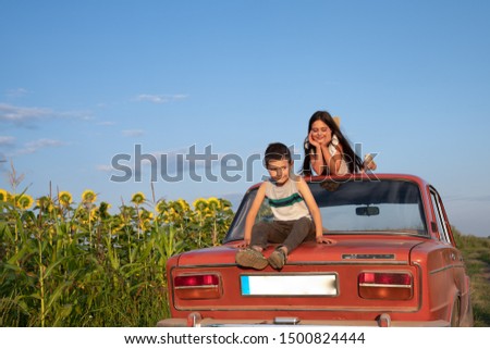 Unsatisfied teen boy in casual clothes sitting on the trunk of the red retro car, her sister is lies on the roof with closed eyes, sunflowers field and blue sky on the background, summer holidays and
