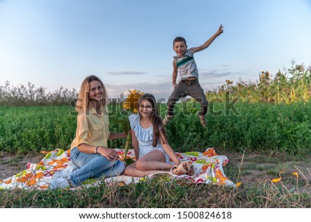 Playful happy family enjoying picnic together , beautiful young mom in yellow shirt sitting on the plaid with her brunette teen daughter in blue striped dress, her little sun is jumping with raised