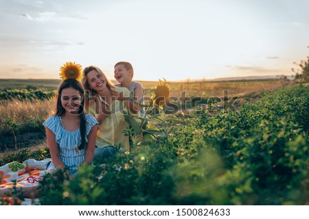 Happy playful family sitting on the plaid among the mealow, brunette teen girl in blue striped dress with sunflower on her head and her mom in yellow shirt with little brother enjoying picnic together
