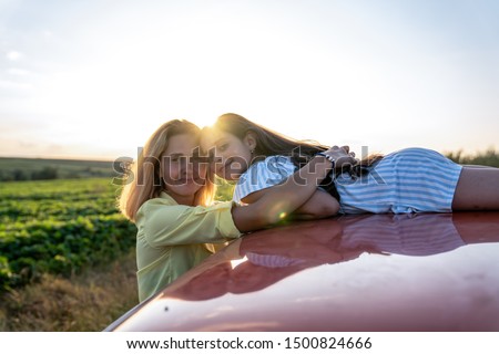 Beautiful smiling family, mom in yellow shirt hugging her dauther in blue striped dress that lies on the roof of the red car, both loooking at the camera, sunset on the background, summer holidays and