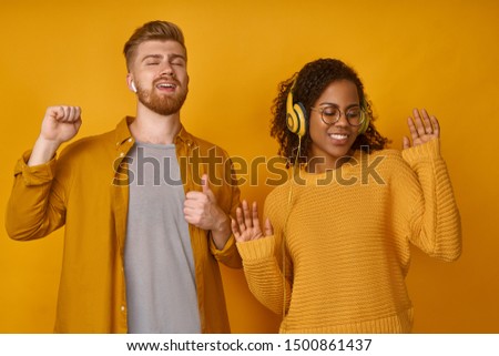 Energized ginger male and black female dance happily, smile broadly, turn at camera, wears headphones, pick right song for good mood, dressed casually, isolated over orange background. Music concept