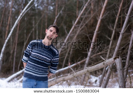 Portrait of a young man tilted towards inclined trees in the background. He is dressed in a sweater and jeans, looking with an empty, sad look. Against the background of a windbreak, snow, forest.