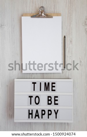 'Time to be happy' words on a lightbox, clipboard with blank sheet of paper on a white wooden background, top view. Overhead, from above. Flat lay. Copy space.