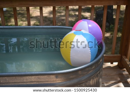 Two colorful, inflatable beach balls float on the water in a stock tank swimming pool on a wooden deck. 