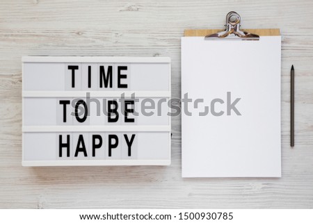 'Time to be happy' words on a lightbox, clipboard with blank sheet of paper on a white wooden background, top view. Overhead, from above. Flat lay. Copy space.
