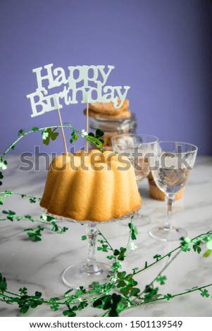 Happy birthday ring cake, cookies, biscuits, muffins and champagne  with clover decoration on marble table and lilac background