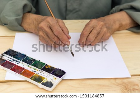 The hand of an elderly man holds a brush for watercolor. The concept of active and happy old age, creativity in adulthood, new beginnings. Image. Royalty-Free Stock Photo #1501164857