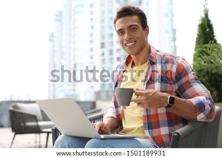 Portrait of handsome young African-American man with laptop and cup of drink in outdoor cafe