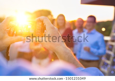 Rear view of man taking pictures of colleagues celebrating sucess at rooftop with yellow lens flare in background