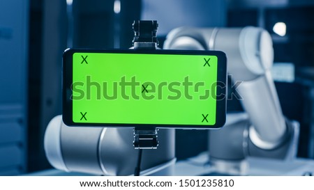 Robot Arm Holding Green Mock-up Screen Smartphone in Landscape Mode. Industrial Robotic Manipulator End Effector Holds Mobile Phone with Chroma Key Display. Royalty-Free Stock Photo #1501235810
