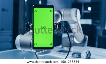 Real Robot Arm Holding Green Mock-Up Screen Smartphone Device. Industrial Robotic Manipulator End Effector Holds Mobile Phone with Chroma Key Display. Modern Manufacturing Facility / Laboratory Royalty-Free Stock Photo #1501235834