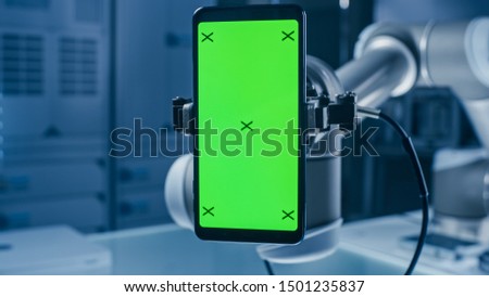 Real Robot Arm Holding Green Mock-Up Screen Smartphone Device. Industrial Robotic Manipulator End Effector Holds Mobile Phone with Chroma Key Display. Modern Manufacturing Facility / Laboratory Royalty-Free Stock Photo #1501235837