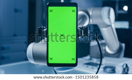 Real Robot Arm Holding Green Mock-Up Screen Smartphone Device. Industrial Robotic Manipulator End Effector Holds Mobile Phone with Chroma Key Display. Modern Manufacturing Facility / Laboratory Royalty-Free Stock Photo #1501235858
