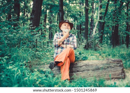 thoughtful man. hiking in deep wood. forest owner. summer or spring picnic. senior man farmer relax in forest. mature man with beard in hat. farmer sit on wood. happy forester. human and nature.