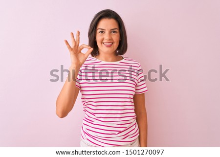 Young beautiful woman wearing striped t-shirt standing over isolated pink background smiling positive doing ok sign with hand and fingers. Successful expression.
