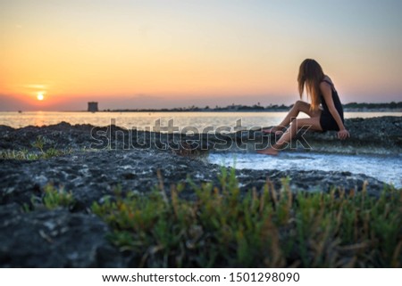 fashion outdoor photo of beautiful woman with dark hair in black mini dress posing and relaxing on summer rock beach. Picture with blurry foreground