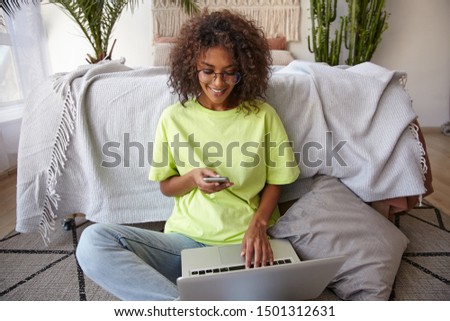 Indoor photo of happy young curly female leaning on bed in sleeping room, working at home with laptop, checking her text messages on her smartphone