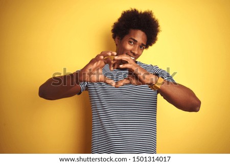African american man with afro hair wearing navy striped t-shirt over isolated yellow background smiling in love showing heart symbol and shape with hands. Romantic concept.